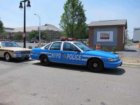 1991 Ford LTD Crown Victoria - blue-and-white NYPD police sedan parks on a street beside a tan classic car, with a Foundry Lofts sign behind.