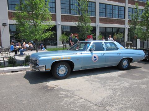 1991 Ford LTD Crown Victoria - light-blue four-door police sedan with a circular door emblem and roof light sits outside a brick building where people gather.