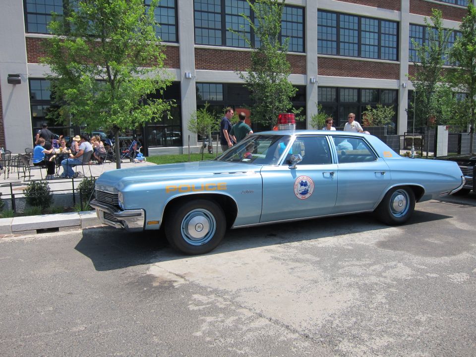 Light blue four-door police sedan with door roundel parked outside brick building