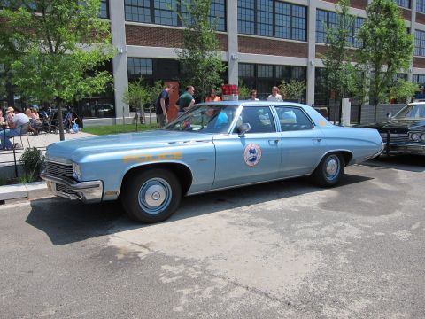 1991 Ford LTD Crown Victoria - light-blue vintage police sedan bearing a circular emblem and roof beacon is parked on pavement outside a brick loft building.