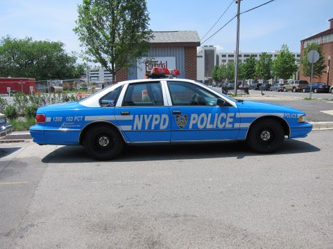 1991 Ford LTD Crown Victoria - bright-blue NYPD Police sedan lettered 1350 103 PCT sits at a curb, twin roof beacons up, before a brick building.