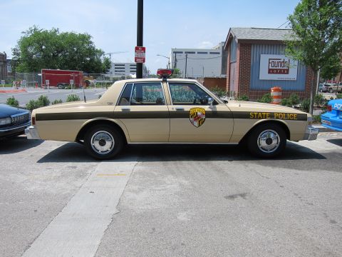 1991 Ford LTD Crown Victoria - tan two-tone State Police sedan bearing a shield door emblem is parked in profile on a street by a brick building.