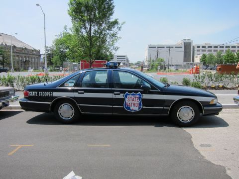 1991 Ford LTD Crown Victoria - black-and-white State Trooper sedan with a State Patrol shield door emblem and roof beacon parks at a curb in an open lot.