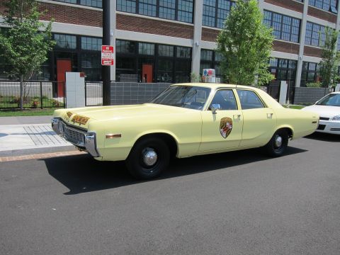 1991 Ford LTD Crown Victoria - pale-yellow police sedan with black wheels and a shield door emblem sits at a three-quarter front angle by a brick loft building.