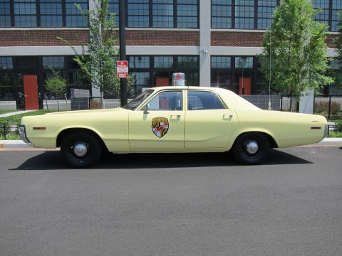 1991 Ford LTD Crown Victoria - pale-yellow vintage police sedan with a shield door emblem and roof beacon is parked in profile along a curb.