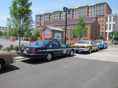 1991 Ford LTD Crown Victoria - dark-blue State Trooper sedan with a shield emblem parks alongside two-tone police cars in a lot before a large brick building.