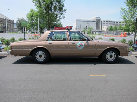 1991 Ford LTD Crown Victoria - tan-brown police sedan with a circular door emblem and roof beacon is parked in profile on pavement under blue sky.