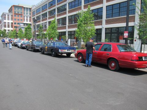 1991 Ford LTD Crown Victoria - line of classic and modern police cars stretches along a curb, ending with a red sedan beside which a man in black stands.