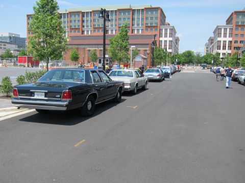 1991 Ford LTD Crown Victoria - black Ford sedan leads a line of vintage sedans parked along a curb beside brick mid-rise buildings.