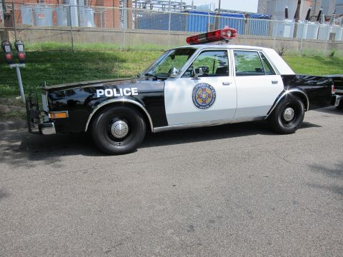 1991 Ford LTD Crown Victoria - Side view of a black-and-white vintage Police sedan with a round agency seal and red roof beacon.