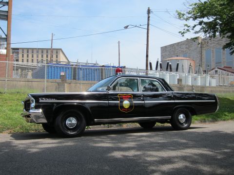 1991 Ford LTD Crown Victoria - Side view of a black 1960s Pontiac police sedan with a gold door shield and single red roof beacon.