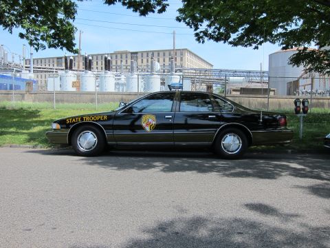 1991 Ford LTD Crown Victoria - black Chevrolet Caprice State Trooper sedan with a Maryland shield decal parked near tanks.