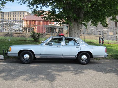 1991 Ford LTD Crown Victoria - light blue full-size four-door sedan with a single red roof beacon sits curbside in profile beside an industrial chain-link fence.