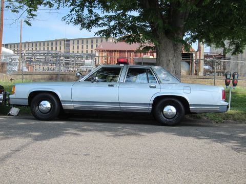 1991 Ford LTD Crown Victoria - light blue full-size four-door sedan with a roof-mounted beacon is parked in profile under a leafy tree near a fenced lot.