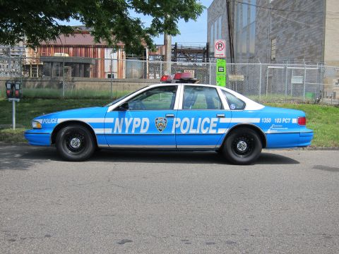 1991 Ford LTD Crown Victoria - blue and white police sedan marked NYPD POLICE 1350 103 PCT sits in profile beside a chain-link fence and No Parking sign.