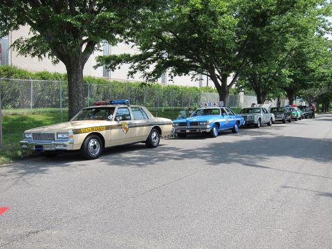 1991 Ford LTD Crown Victoria - line of vintage police cars including a tan STATE POLICE sedan and a blue and white cruiser parked along a shaded tree-lined street.