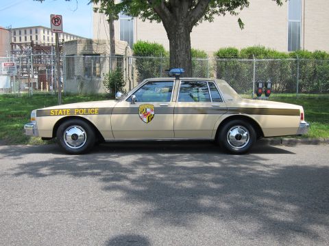 1991 Ford LTD Crown Victoria - tan four-door sedan marked STATE POLICE with a Maryland crest and single roof beacon sits in profile beside a chain-link fence.