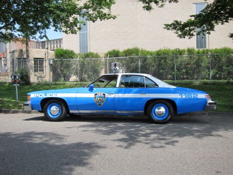 1991 Ford LTD Crown Victoria - blue and white two-door vintage police car numbered 1162 with a roof beacon sits in profile on a sunlit street by a fence.