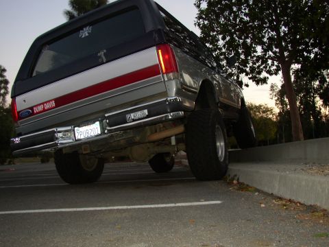 1989 Ford Bronco - This lifted Ford Bronco is parked with its rear wheels on a curb, showing off its large tires and aftermarket modifications.