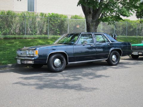 1991 Ford LTD Crown Victoria - dark blue full-size four-door sedan is parked in front three-quarter view beside a chain-link fence and tree.