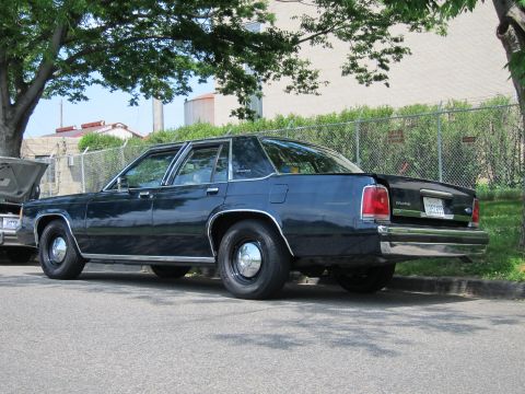 1991 Ford LTD Crown Victoria - dark blue full-size four-door sedan is shown in rear three-quarter view parked along a curb beside a fenced industrial lot.