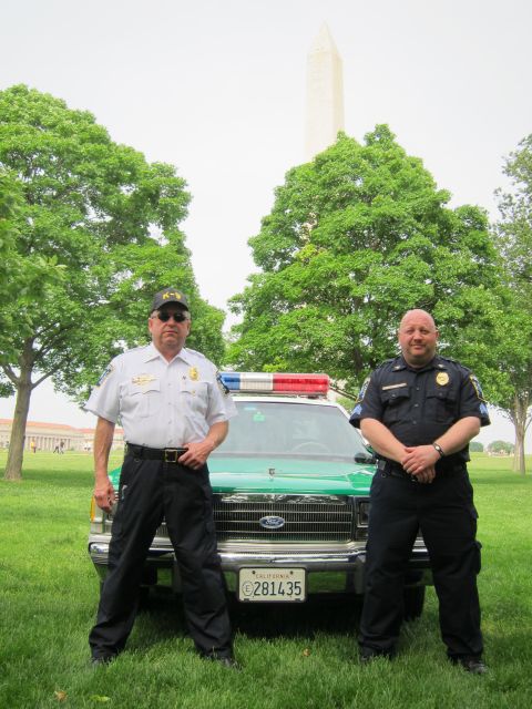 1991 Ford LTD Crown Victoria - Two officers stand in front of a green-and-white police cruiser on a lawn with the Washington Monument framed by trees behind them.
