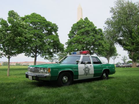 1991 Ford LTD Crown Victoria - green-and-white police sedan with a roof lightbar sits on a grassy lawn, the Washington Monument rising behind the trees.