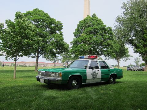 1991 Ford LTD Crown Victoria - green-and-white police cruiser with a roof lightbar sits on a grassy field beneath trees with the Washington Monument in view.