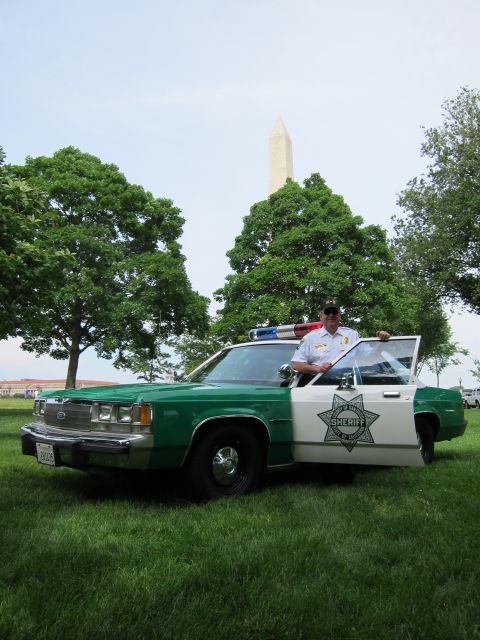 1991 Ford LTD Crown Victoria - officer stands in the open door of a green-and-white Sheriff sedan with a star emblem, the Washington Monument rising behind.
