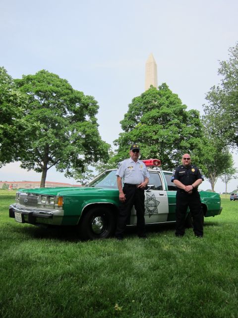 1991 Ford LTD Crown Victoria - Two uniformed officers pose in front of a green-and-white police sedan with a roof lightbar and the Washington Monument behind.