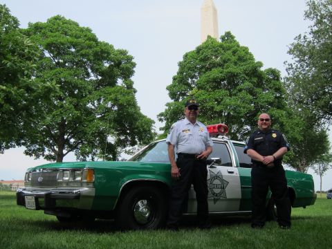 1991 Ford LTD Crown Victoria - Two officers stand beside a green-and-white police cruiser bearing a star door emblem with the Washington Monument behind on a lawn.