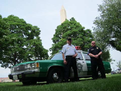 1991 Ford LTD Crown Victoria - Two officers pose beside a green-and-white police sedan with a roof lightbar, the Washington Monument framed by trees behind them.
