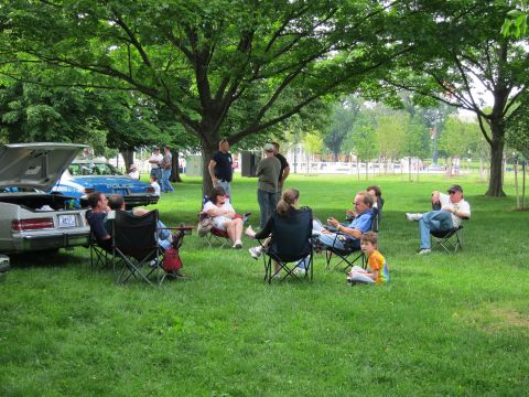 1991 Ford LTD Crown Victoria - group of people relax in folding camp chairs on a shaded lawn beside a parked car with its trunk open.
