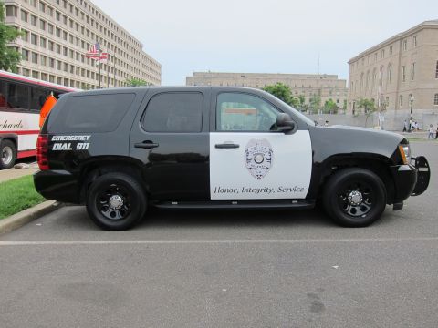 1991 Ford LTD Crown Victoria - black-and-white Chevrolet police SUV displaying 'Emergency Dial 911' and a badge logo is parked along a city curb.