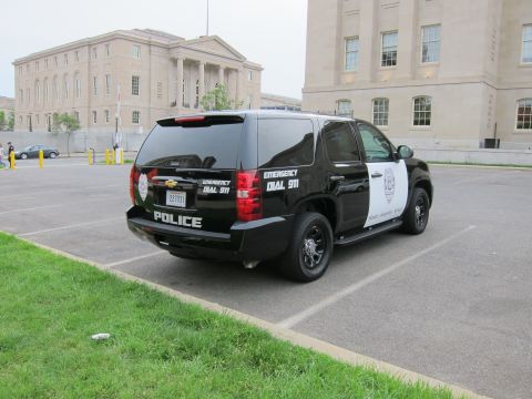 1991 Ford LTD Crown Victoria - black-and-white Chevrolet police SUV marked 'POLICE' and 'Emergency Dial 911' is parked in a lot before stone buildings.