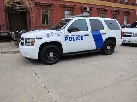 1991 Ford LTD Crown Victoria - white-and-blue Chevrolet SUV marked 'Federal Protective Service Police' with a Homeland Security seal parked by a brick building.