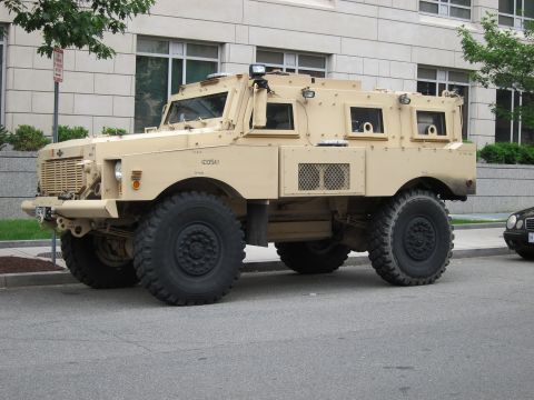 1991 Ford LTD Crown Victoria - tan armored MRAP-style military vehicle with large tires and a winch is parked along a city street beside a stone building.