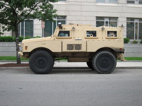 1991 Ford LTD Crown Victoria - tan armored MRAP military vehicle with large off-road tires parked along a city street beside a stone building.
