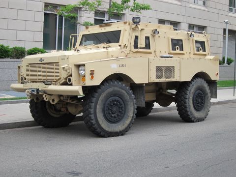 1991 Ford LTD Crown Victoria - Front three-quarter view of a tan armored MRAP vehicle with a winch and heavy off-road tires parked curbside.