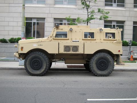 1991 Ford LTD Crown Victoria - Side profile of a tan armored MRAP military vehicle with armored windows parked on an urban street.