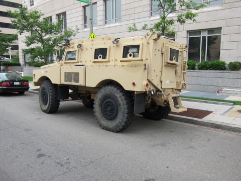 1991 Ford LTD Crown Victoria - Tan armored MRAP military vehicle viewed from the rear quarter, parked near a building with a sedan behind it.