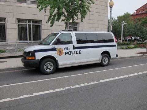 1991 Ford LTD Crown Victoria - white FBI Police passenger van with blue striping and gold shield, parked along an urban curb.