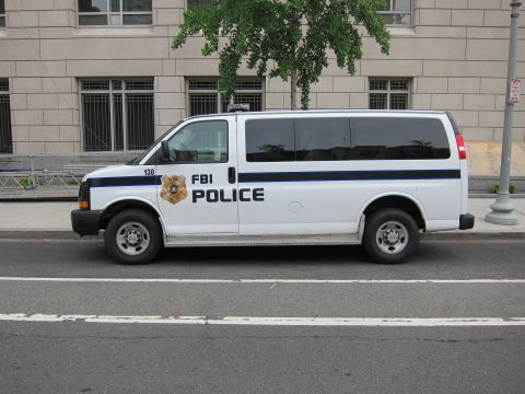 1991 Ford LTD Crown Victoria - Side profile of a white FBI Police van marked with a gold shield and blue stripes parked on a city street.