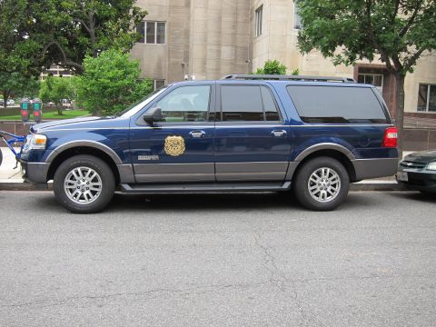 1991 Ford LTD Crown Victoria - dark blue Ford Expedition with a gold federal agency shield on the door, parked along a city street.