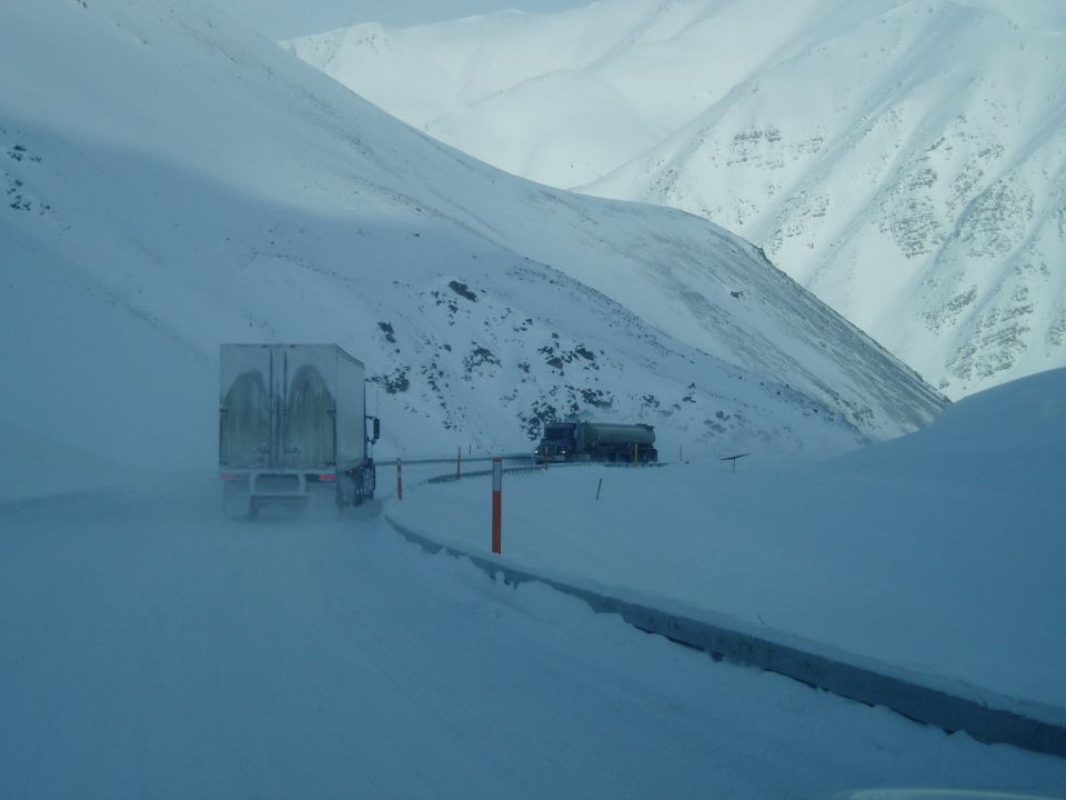 Two trucks driving on a snow-covered mountain road with steep, snow-covered slopes