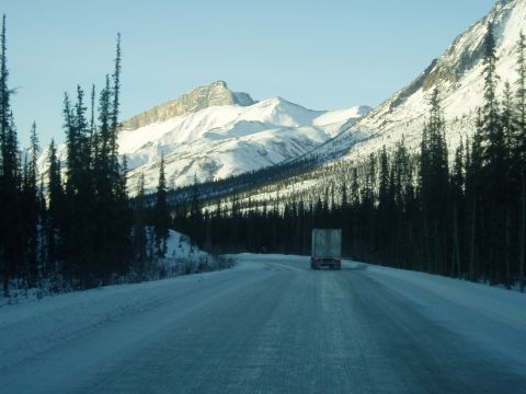 2009 Kenworth T800 - A semi-truck travels down a snowy mountain highway surrounded by dark evergreen trees and snow-capped peaks.