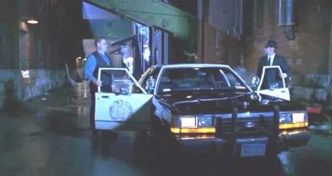 1991 Ford LTD Crown Victoria - A police car is parked in an alleyway with two men standing by its open doors.
