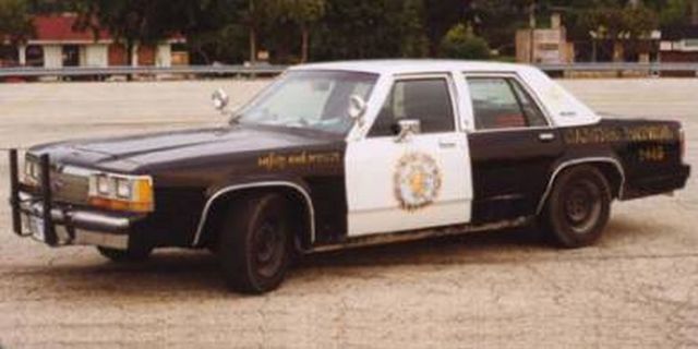 Black and white Ford police sedan with push bar and pillar spotlights on a dirt lot