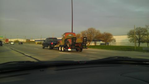 1989 Ford Bronco - An old truck is transported on a trailer behind a dark SUV on a highway under a cloudy sky.