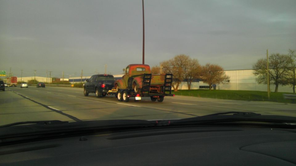 Old truck on a trailer being towed by a dark SUV on a highway